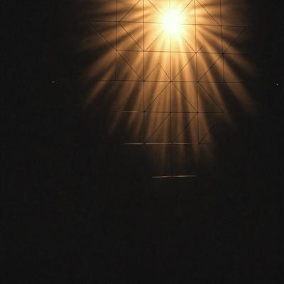The primary subject of the image is a bright light source, possibly a sun or a light bulb, shining through a patterned surface. The setting appears to be a dark room, with the light source illuminating the patterned surface. The visual style of the image is a combination of a photo and a digital art, as it captures the light source's reflection on the patterned surface. The colors and mood of the image are predominantly dark, with the bright light source creating a contrasting and visually striking